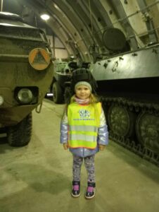 Dziewczynka w hełmofonie stojąca obok czołgu.